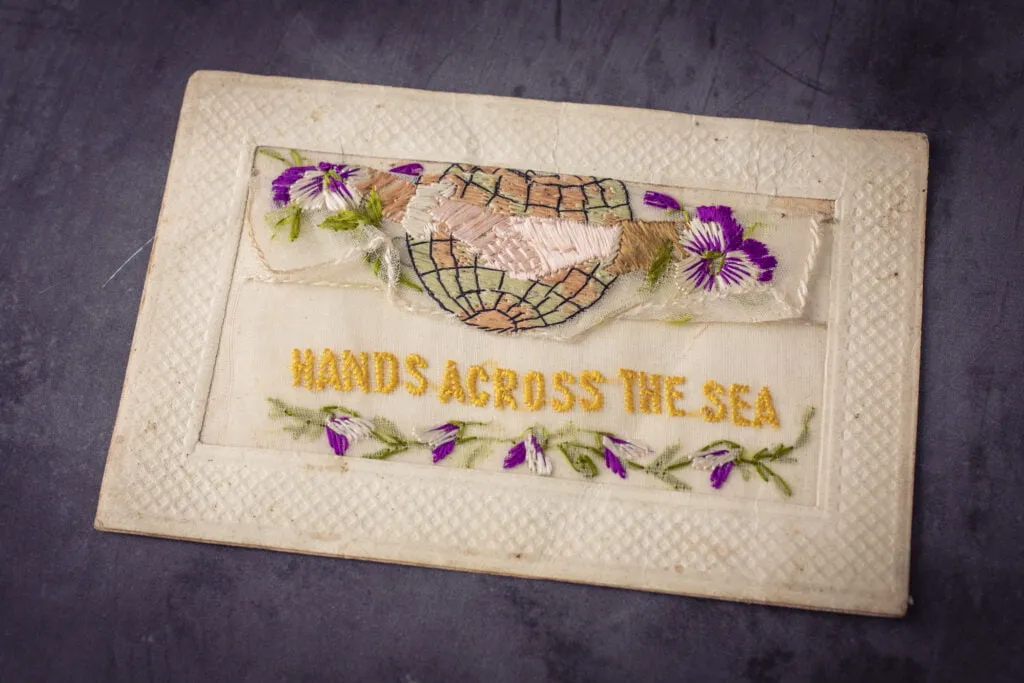 Vintage embroidery war postcard on lace of flowers and two hands shaking in front of the globe with the script hands across the sea underneath 