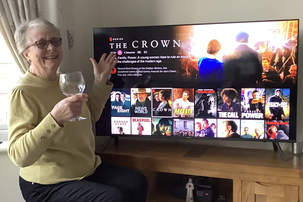 happy woman with a drink in her hand and a TV with The Crown on in the background