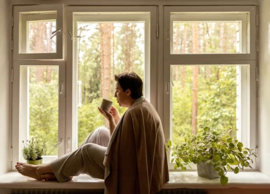 woman with blanket blows a cup of tea while looking out of window